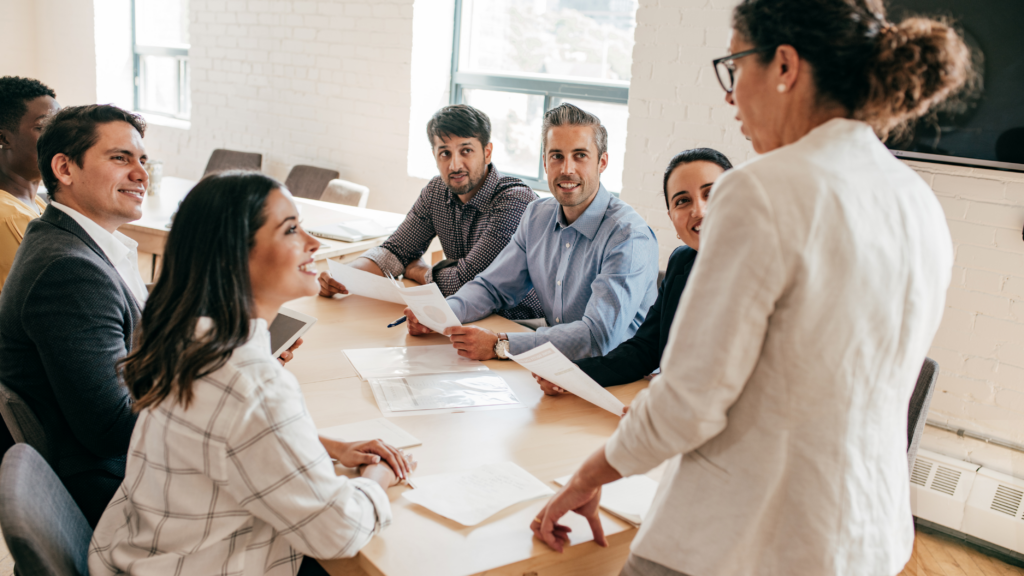 Leading Teams Through Change - A woman standing, addresses her seated team as she coaches and encourages them through a period of change.