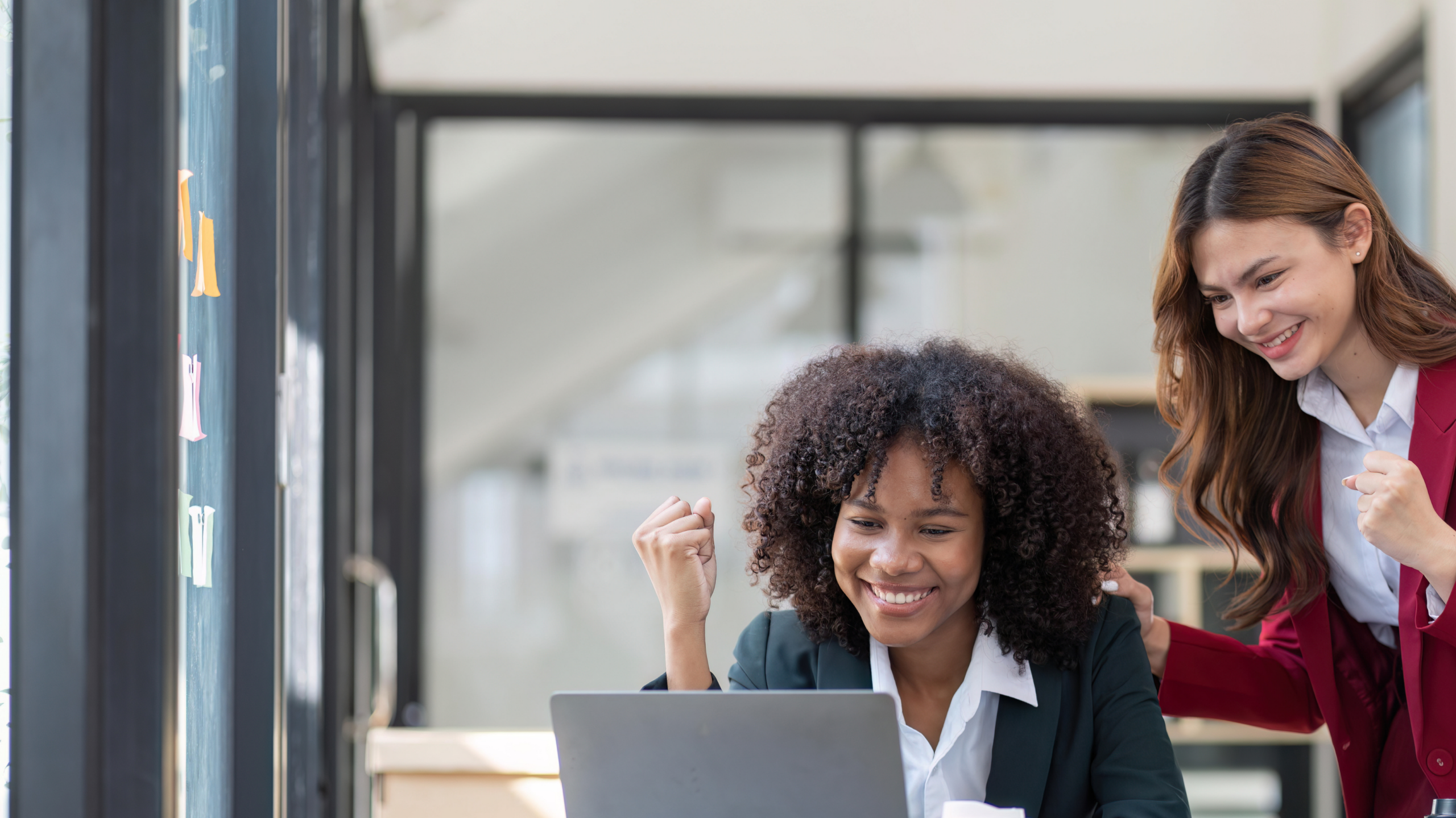 Two smart young women look at a laptop screen in an office, looking jubilant as they celebrate managing performance for success.